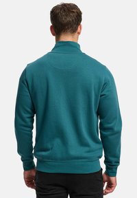 Blågrønn genser med høy krage, ribbestrikkede mansjetter og linning. Jevn tekstur, minimalt med detaljer, og en tilpasset snitt bak.