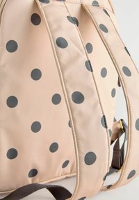 Zaino beige con pois neri. Presenta spallacci imbottiti e una texture di tessuto liscia. I dettagli includono una striscia a contrasto e accessori in metallo.