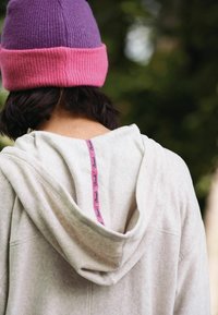Personne portant un bonnet violet et rose et un sweat à capuche gris clair avec une étiquette rose visible à l'arrière de la capuche.