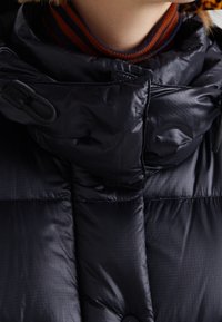 Close-up af en sort polstret jakke med en aftagelig rem på kraven, båret over en brun og marineblå stribet højhalset trøje.