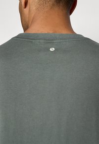 Zaļš kokvilnas T-krekls ar apaļu kakla izgriezumu un mazu logotipa detaļu aizmugurē. Audumam ir gluda, nedaudz teksturēta virsma.