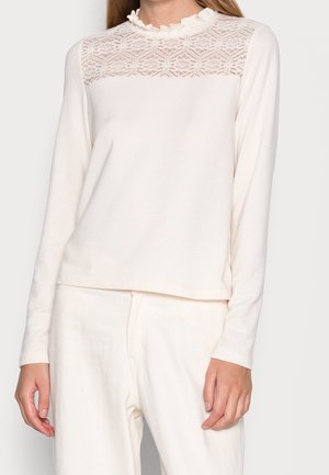 Haut à manches longues couleur crème avec un empiècement en dentelle et un col à volants. Confectionné dans un tissu doux, il présente une coupe décontractée et une texture lisse.
