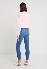 Růžová mikina s kulatým výstřihem, přiléhavé modré džíny a bílé nazouvací boty. Oblečení má ležérní, uvolněný střih a ohrnuté manžety.