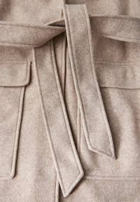 Manteau en laine beige avec deux poches avant, une ceinture nouée et des détails cousus sur les bords, avec une finition douce et texturée.