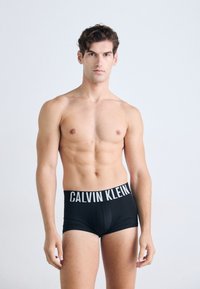 Schwarze Calvin Klein Boxershorts mit einem weißen Markenbund. Das Model hat einen definierten Oberkörper und steht vor einem neutralen Hintergrund.