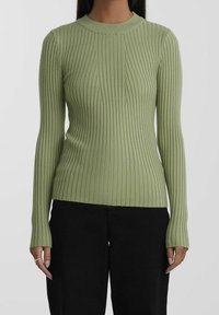 Femme portant un pull vert clair à manches longues côtelé avec un pantalon noir, montrant le haut du corps et les bras sur un fond uni.