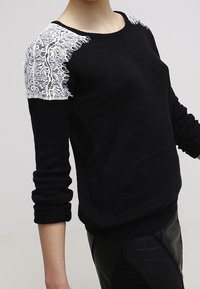 Pull en maille noire avec panneaux d'épaule en dentelle blanche, porté par une personne aux manches retroussées, sur un fond uni.