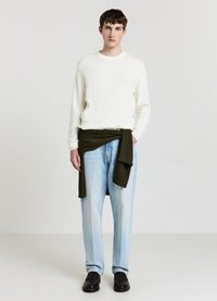 Maglione bianco a coste, jeans larghi azzurro chiaro, maglione verde lavorato a maglia annodato in vita, mocassini neri, sfondo semplice.