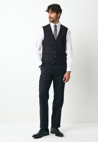 Gilet noir à cinq boutons porté sur une chemise blanche, associé à un pantalon noir et des chaussures noires cirées. Texture lisse semblable à de la laine.