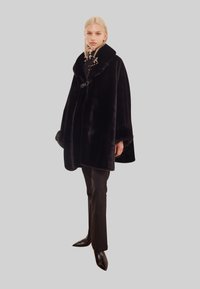 Svart fuskpäls-cape med bred krage, med en knappstängning och texturerade detaljer. Bärs med figurnära svarta byxor och spetsiga skor.