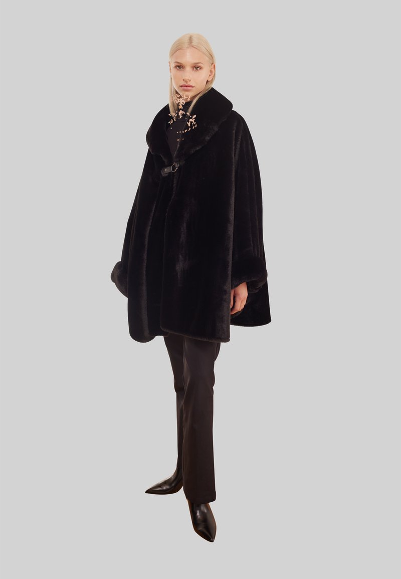 Svart fuskpäls-cape med bred krage, med en knappstängning och texturerade detaljer. Bärs med figurnära svarta byxor och spetsiga skor.