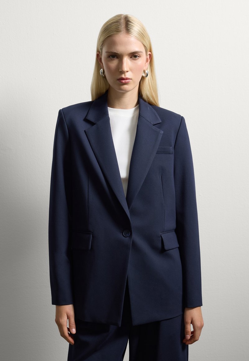 Blazer sartorial blu navy con spalle strutturate, due tasche frontali, chiusura a un bottone e superficie in tessuto liscia.