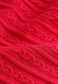 Roter gestrickter Stoff mit einem strukturierten Kabelmuster, das erhabene Muster und eine weiche, flauschige Textur aufweist, die in wellenförmigen Falten angeordnet ist.
