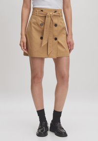 Jupe en coton de couleur camel, arrivant au genou. Elle présente une taille ceinturée, un design à double boutonnage avec des boutons noirs, et une coupe droite.