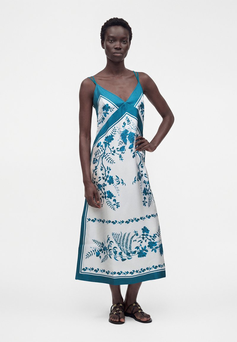 Woman wearing a teal and white floral midi dress with thin straps, standing with one hand on hip and black sandals on a plain background.
