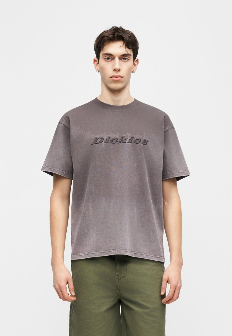 Giovane uomo con capelli corti e scuri, indossa una maglietta grigia sbiadita Dickies e pantaloni verde oliva, in piedi davanti a uno sfondo bianco uniforme.