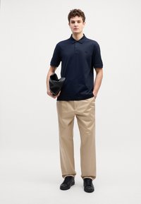 Pólo azul-marinho com colarinho de três botões, calças bege e ténis pretos, segurando uma bolsa preta. Design simples, textura de tecido suave.