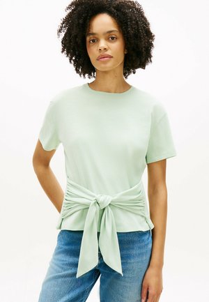 Frau mit lockigem Haar, die ein hellgrünes Kurzarmhemd trägt, das vorn zu einem Knoten gebunden ist, und blaue Jeans, steht vor einem weißen Hintergrund.