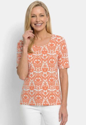 Lächelnde Frau mit langen blonden Haaren, die ein kurzärmeliges, orange-weißes, floral gemustertes Oberteil und weiße Hosen trägt, steht vor weißem Hintergrund.