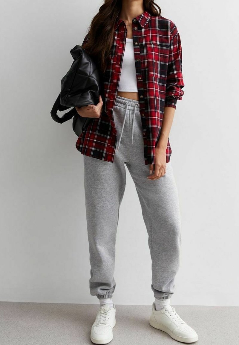 Femme portant une chemise rouge à carreaux par-dessus un crop top blanc, un pantalon de survêtement gris, des baskets blanches, et tenant un sac à dos noir sur un fond uni.