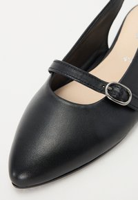 Chaussure en cuir noir avec un bout pointu, une fine lanière ornée d'une boucle ronde en argent et un dessus à texture lisse.