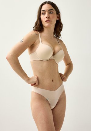 Kvinde med langt brunt hår iført beige bh og matchende undertøj, stående med hænderne på hofterne og kiggende til siden mod en ensfarvet baggrund.
