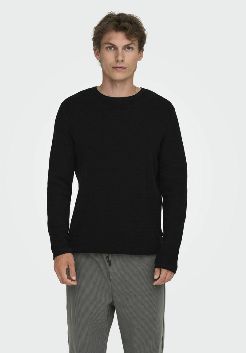 Schwarzer Pullover mit langen Ärmeln und Rundhalsausschnitt, aus strukturiertem Strickmaterial mit enganliegenden Bündchen, kombiniert mit olivgrünen Cordhosen.