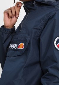 Marinblå vindjacka med halvmatch, elastiska ärmslut och en framficka. Har logotypmärken i orange, vitt och rött på bröstet och ärmen.