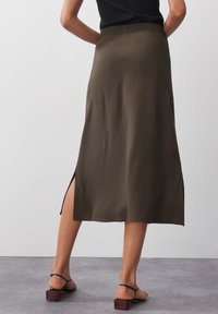 Oliivinvihreä midihame, jossa on sileä pinta, joustava vyötärönauha ja sivuhalkiot, yhdistetty mustiin remmihinnoilla varustettuihin sandaaleihin.