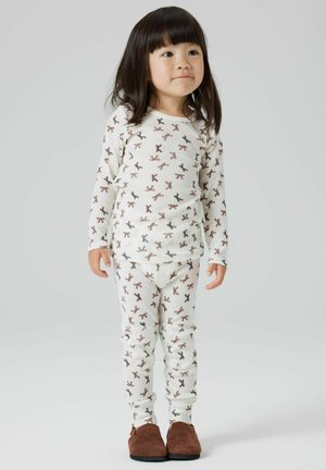 Ensemble de pyjama en coton blanc avec des motifs répétés de nœuds marron, manches longues et pantalon ajusté. Le modèle porte des chaussures marron.