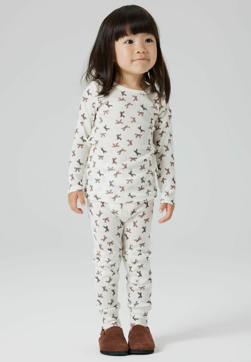 Ensemble de pyjama en coton blanc avec des motifs répétés de nœuds marron, manches longues et pantalon ajusté. Le modèle porte des chaussures marron.
