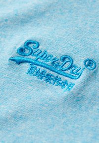 Tessuto in cotone azzurro chiaro con un logo ricamato "Superdry" in un blu più scuro, caratterizzato da una texture liscia e senza pattern visibili.