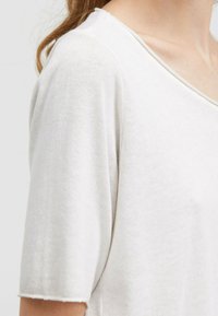 Helles beige Oberteil mit kurzen Ärmeln und V-Ausschnitt. Hergestellt aus weichem, strukturiertem Stoff, mit einem leicht lockeren Schnitt und unvollendeten Ärmelrändern.