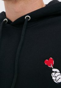 Černá mikina z jemného materiálu s měkkou texturou, obsahující bílou grafiku ruky, která drží červené srdce, doplněná vyšitými detaily a oky.