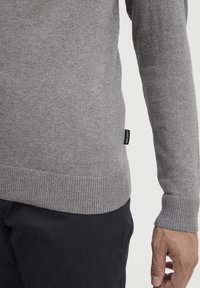 Grauer Pullover aus weichem Stoff mit geripptem Saum und Bündchen. Verfügt über ein kleines Logoschild an der Seite und ein tailliertes Design.