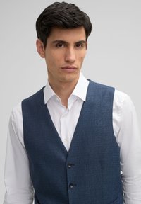 Navyblaues, strukturiertes Weste aus Stoff mit drei Knöpfen, kombiniert mit einem weißen Hemd mit Kragen. Figurbetontes Design, keine sichtbaren Muster oder Akzente.
