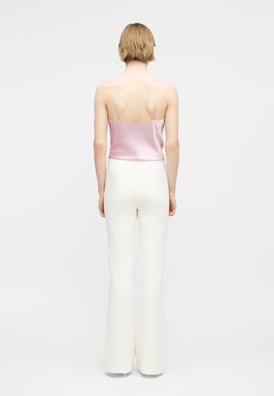 Γυναίκα με κοντά ξανθά μαλλιά που φοράει ροζ σατέν τοπ και λευκό παντελόνι με φαρδιά μάτια και ψηλή μέση, στέκεται έχοντας την πλάτη της προς το φακό σε λευκό φόντο.