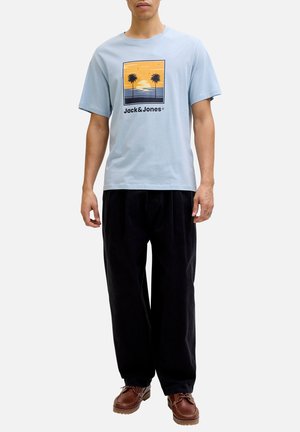 Homme portant un t-shirt bleu clair avec un graphique coucher de soleil palmier et le texte « Jack & Jones », pantalon noir et chaussures marron, debout devant un fond blanc.