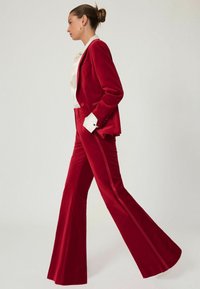 Červený sametový oblek so priliehavým sako a rozšírenými nohavicami. Obsahuje bielu košeľu s volánovým golierom a kontrastnými čiernymi manžetami.