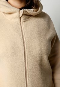 Chaqueta de forro polar beige con una superficie texturizada, cierre completo y capucha. La tela parece suave y cálida, con un ajuste general oversize.