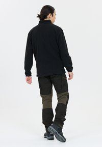 Zwarte fleecepullover met een hoge kraag, gedragen met donker groene en zwarte multifunctionele broek met meerdere zakken en zwarte wandelschoenen, gezien van achteren.