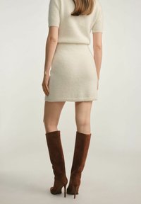 Kremowa dzianinowa sukienka z krótkimi rękawami, dopasowany fason i ściągany dół, w połączeniu z wysokimi brązowymi zamszowymi botkami na szpilce, widok z tyłu.