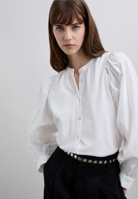 Blusa blanca abotonada con mangas abullonadas, con un escote redondeado y tejido de algodón suave y texturizado. Combinada con pantalones negros de cintura alta.