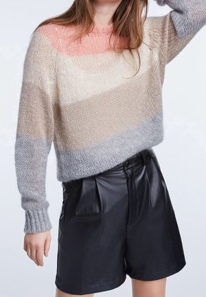 Kobieta ubrana w miękki, pasiasty sweter w kolorach różowym, beżowym i szarym, w połączeniu z czarnymi skórzanymi szortami z wysokim stanem, pozująca z uniesioną jedną ręką.