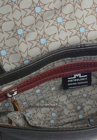 Interno di una borsa in pelle nera con una fodera a fantasia color marrone chiaro con quadrati blu e forme di stelle. Include una tasca con zip color bordeaux.