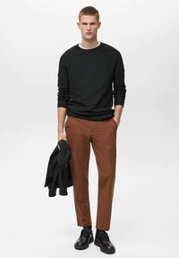 Črn pulover s krojnim ovratnikom, rjave hlače in črne čevlje. Model drži črno jakno in stoji ob svetli enotni ozadju.