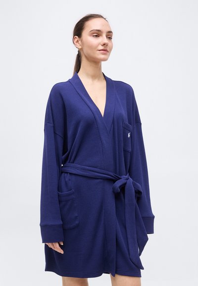 Νεαρή γυναίκα που φοράει μια μπλε ναυτική ρόμπα με ζώνη, μακριά μανίκια και δύο μπροστινές τσέπες, με απλό ανοιχτό φόντο.