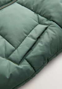 Gros plan d'une veste matelassée verte avec une poche horizontale cousue et une texture de tissu visible.