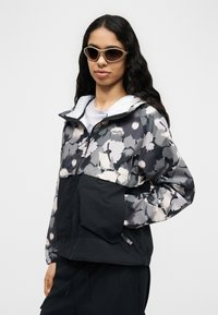 Γυναίκα με μακριά σκούρα μαλλιά φοράει γκρι και μαύρο μπουφάν με λουλουδάτο σχέδιο και μπεζ γυαλιά ηλίου, στέκεται με το ένα χέρι στην τσέπη.