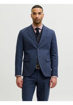 Mann iført en marineblå dress med matchende vest, lyserosa skjorte og svart slips, stående foran en ensfarget lys bakgrunn.